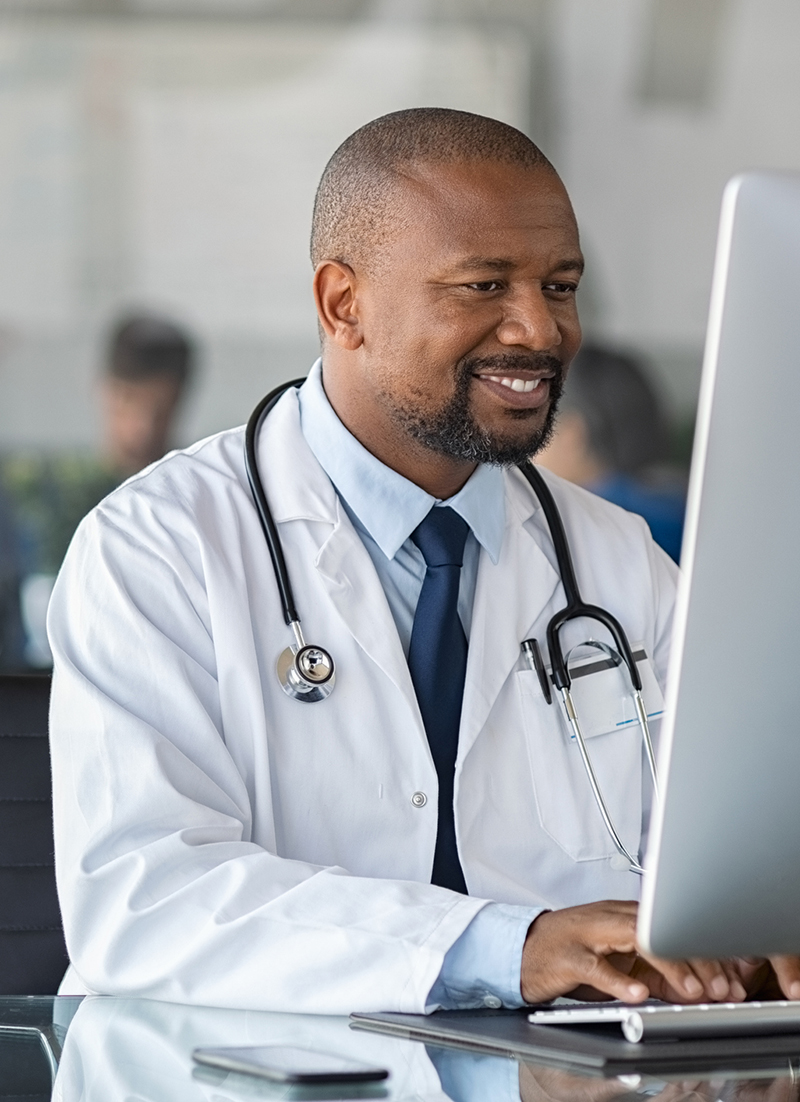 Practitioner typing on computer in modern clinic. Doctor at work in office using computer with copy space. African happy specialist wearing white coat and sitting at desk while mediacal team having meeting in background.
